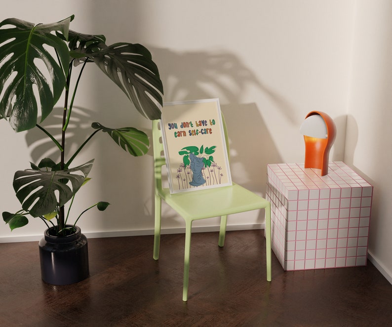 May include: A green chair with a framed print that reads "You don't have to earn self-care" with a cartoon illustration of a plant. The chair is in front of a white wall and a small white and pink tiled table with a lamp on top.