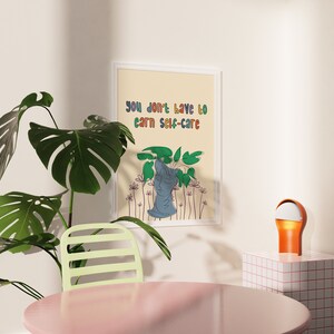 May include: A framed print with a light beige background and a colorful text overlay that reads "you don't have to earn self-care". The print features a stylized illustration of a person with a plant growing out of their head.