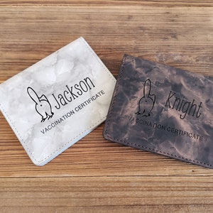 May include: Two leather vaccination certificate holders. One is white with a marble pattern and the name "Jackson". The other is brown with the name "Knight". Both have a graphic and the text "VACCINATION CERTIFICATE".