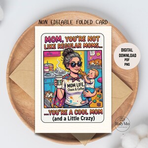 Può includere: Biglietto di auguri piegato con un'illustrazione a cartoni animati di una donna che tiene in braccio un bambino e una tazza di caffè. Il biglietto recita "MOM, YOU'RE NOT LIKE REGULAR MOMS... YOU'RE A COOL MOM (and a Little Crazy)". Il biglietto è su un vassoio di legno.