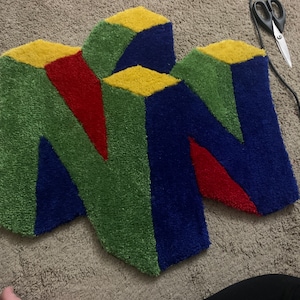 May include: A colorful rug with the Nintendo 64 logo in a 3D design. The rug is made of a soft, plush material and features shades of blue, green, red, and yellow.
