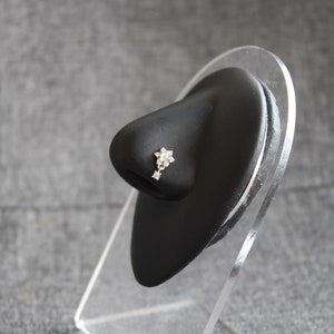 Könnte beinhalten: Ein silberner Nasenring mit einem kleinen Blumenmuster und klaren Strasssteinen. Der Ring wird auf einem schwarzen Ständer präsentiert.