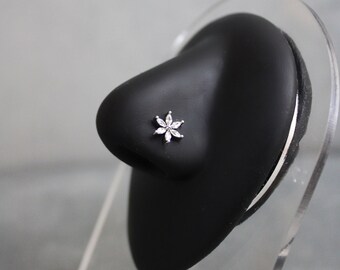 May include: A silver nose stud featuring a flower design with six clear, sparkling petals. The jewelry is set on a black nose form, displayed on a clear acrylic stand. The nose stud is a delicate piece of body jewelry.