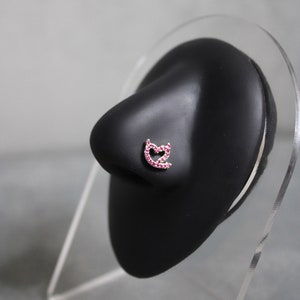 Puede incluir: Un aro de nariz de plata con un diseño en forma de corazón, engastado con piedras preciosas rosas.