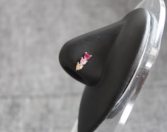 May include: A silver nose ring with three heart-shaped gemstones. The gemstones are pink, peach, and clear.