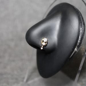 May include: Gold nose ring with three clear cubic zirconia stones. The ring is on a black display stand.