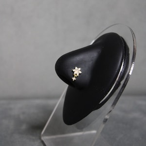 Puede incluir: Un piercing de nariz dorado con un diseño floral, una piedra central similar al ópalo y detalles colgantes. La joya se muestra sobre un modelo de nariz negra sobre un fondo gris.