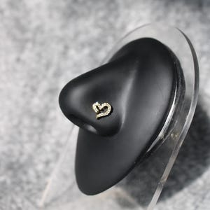 May include: A gold-coloured nose stud shaped like a heart, set with small, clear gemstones. The stud is displayed on a black nose form against a grey background. The jewellery is designed for nose piercings.
