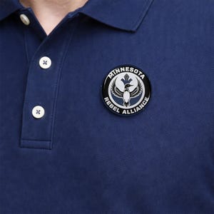 May include: Navy blue polo shirt with a white collar and buttons. A circular emblem with the text "MINNESOTA REBEL ALLIANCE" surrounds a stylized bird graphic. The shirt has a classic, casual design.