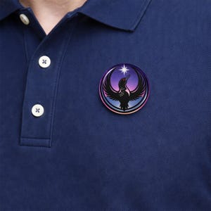 May include: A round, colorful pin featuring a black bird with outstretched wings, a red eye, and a star. The background transitions from purple to blue. The pin is attached to a navy blue polo shirt.