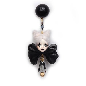 May include: A black and white doll head with a black bow and a black balloon attached to the top. The doll has white hair and black makeup around the eyes. The doll is wearing a black bow and has a gold star charm hanging from the bottom.