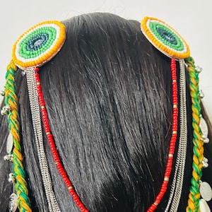 May include: A colorful headpiece featuring a braided green and yellow cord, adorned with silver coins, cowrie shells, and dangling chains. Two circular accents with green and orange embroidery are attached to the hair. A red beaded strand adds a pop of color.