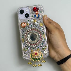 May include: Clear phone case with intricate embellishments. The case features a central mirror surrounded by silver beads and colorful gems. A gold pendant with green and red accents hangs at the bottom, adding a touch of elegance.