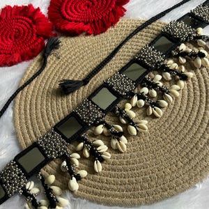 Puede incluir: Un collar negro estilo cinturón con detalles de espejo cuadrados, cuentas plateadas y conchas blancas colgantes. También hay dos posavasos rojos circulares con flecos y borlas negras.