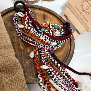 May include: A hair accessory featuring multiple braided cords in black, white, red, and brown. The strands are adorned with white cowrie shells, small turquoise beads, and a single red wooden bead, all secured by a black elastic hair tie.