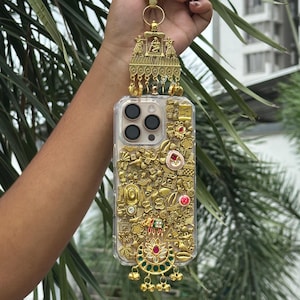 May include: A clear phone case decorated with gold-coloured embellishments and a matching gold-coloured charm. The case features various shapes and designs, including a central pendant with red and green accents. The charm has a decorative top with colourful stones.