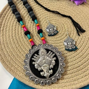 Puede incluir: Un conjunto de collar y pendientes plateados. El collar presenta un colgante grande con una figura central, rodeado por un borde decorativo. El collar tiene un cordón negro con cuentas de colores. Los pendientes tienen un diseño similar con una borla turquesa.