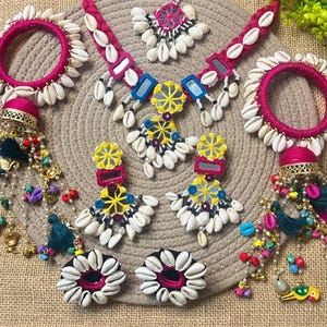 May include: A jewelry set featuring a pink thread-wrapped necklace with cowrie shells and mirrors, two matching pink bangles with cowrie shells, two yellow and pink mirror earrings, and two dangling hair accessories with colorful beads and charms.