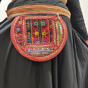 Puede incluir: Un cinturón con un bolsillo bordado rojo y circular. El bolsillo presenta intrincados patrones en rojo, verde, azul y amarillo. El cinturón es negro con una banda estampada. El cinturón se lleva sobre una falda negra.