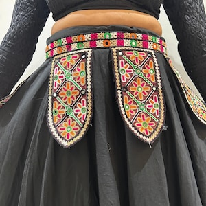 Könnte beinhalten: Schwarzer Rock mit farbenfrohen Stickereidetails. Der Rock hat einen breiten Bund mit einem lebendigen Muster aus Rosa, Grün, Orange und Weiß. Der Rock hat dekorative Einsätze mit Blumenstickereien in ähnlichen Farben. Der Rock ist aus fließendem Stoff.