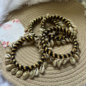 May include: Three black and gold bangles adorned with cowrie shells. The bangles have a striped design and are accented with small gold beads. The cowrie shells are cream-colored and hang from the bangles.