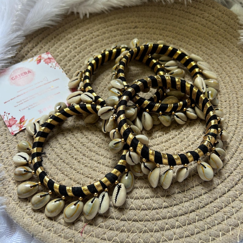 May include: Three black and gold bangles adorned with cowrie shells. The bangles have a striped design and are accented with small gold beads. The cowrie shells are cream-colored and hang from the bangles.