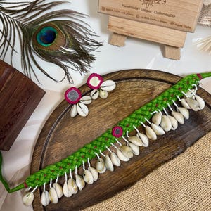 May include: A handmade green choker necklace with white seashells and pink accents. Matching seashell earrings are also present. The jewelry is displayed on a wooden tray with a peacock feather and a wooden box in the background.