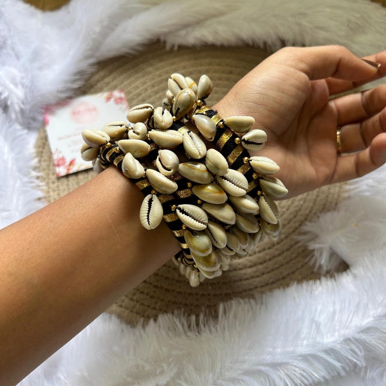 May include: A stack of bracelets adorned with numerous cream-colored seashells. The bracelets feature a black and gold striped pattern. The seashells are clustered together, creating a textured and eye-catching accessory.