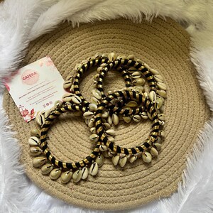 May include: A set of three black and gold bracelets adorned with numerous white seashells. The bracelets are arranged on a woven, circular mat, with a business card in the background. The card has the name "GAJERA" printed on it.