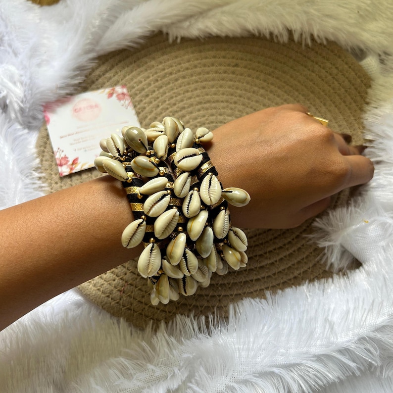 May include: A wide bracelet featuring numerous cream-colored seashells. The bracelet has a black band with gold stripes and is adorned with small gold beads. The bracelet is displayed on a wrist.