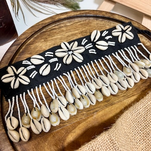 Puede incluir: Una gargantilla de tela negra con conchas de cauri blancas dispuestas en patrones florales. La pieza está adornada con flecos de cuentas blancas colgantes, con conchas de cauri adicionales unidas a los extremos para un aspecto texturizado.