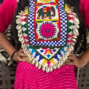 May include: A white top with colorful embroidery featuring an elephant, flowers, and geometric patterns. The top has a scalloped neckline and is adorned with white shell beads.