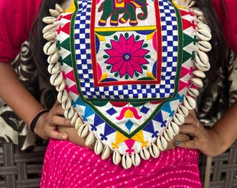 May include: A white top with colorful embroidery featuring an elephant, flowers, and geometric patterns. The top has a scalloped neckline and is adorned with white shell beads.