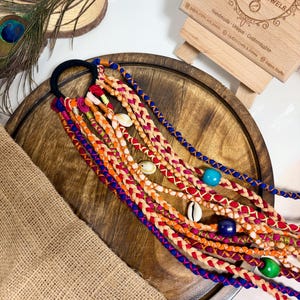May include: A hair accessory featuring multiple braided strands of colorful yarn in shades of orange, red, blue, and pink attached to a black elastic band. The braids are decorated with white cowrie shells and solid blue and green beads.