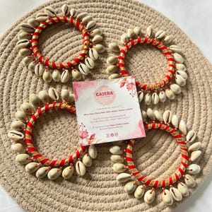 May include: Four handcrafted bracelets with red and gold accents, adorned with white seashells. The bracelets are arranged on a woven mat, with a small card featuring the brand name "GAJERA" and floral designs.