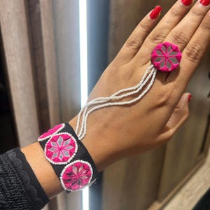 Puede incluir: Un conjunto de pulsera y anillo negros con diseños florales rosas y plateados. La pulsera tiene tres acentos bordados circulares. El anillo está conectado a la pulsera con tres hebras de cuentas blancas. La joyería está adornada con pequeños espejos.