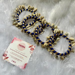 May include: Three blue and gold bangles adorned with cream-colored seashells. The bangles are arranged on a white, textured surface. A small card with the text "GAJERA" and floral accents is visible.