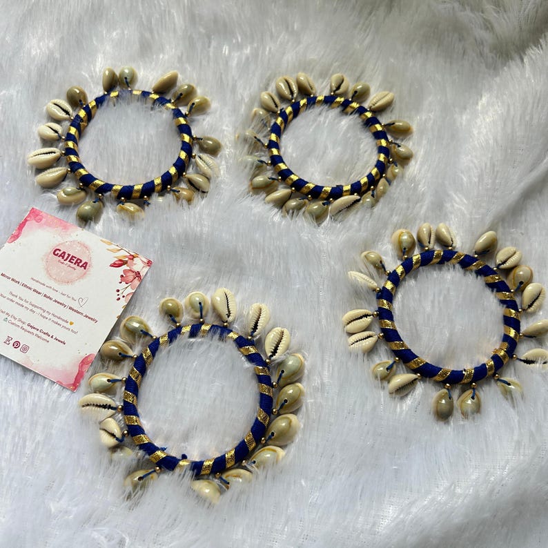 May include: Four blue and gold bracelets adorned with cream-colored seashells. The bracelets are arranged on a white, fluffy surface. A business card with the name "GAJERA" is visible in the image.