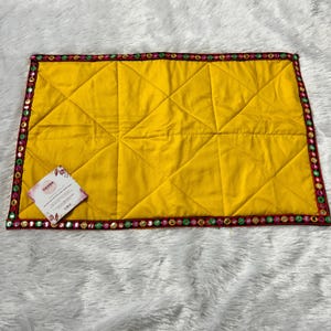 Könnte beinhalten: Ein rechteckiger, gelber Tischset mit einem Steppmuster. Der Rand ist mit kleinen, farbigen Spiegelakzenten verziert. Ein kleines Etikett mit Text befindet sich in der linken unteren Ecke. Das Tischset liegt auf einer weißen, strukturierten Oberfläche.