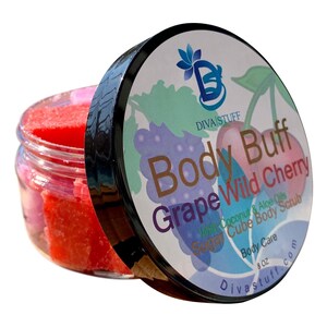 Puede incluir: Un tarro de exfoliante corporal rojo con la tapa ligeramente entreabierta. La tapa presenta las palabras "Body Buff Grape Wild Cherry" con ilustraciones de uvas y cerezas. El tarro contiene 227g de exfoliante.