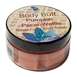 Puede incluir: Un tarro transparente de exfoliante corporal de azúcar en cubos con tapa negra. La etiqueta dice "Body Buff Pumpkin Pecan Waffle Sugar Cube Body Scrub" y "8 OZ Body Care". Los cubos exfoliantes son de color naranja claro.