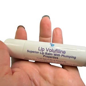 May include: A white cylindrical lip balm tube labeled Lip Volufiline Superior Lip Balm With Plumping Properties. The label includes a small blue logo, usage instructions, and a list of ingredients printed in dark blue text on the white surface.