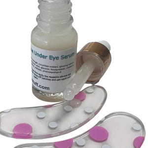 May include: A clear glass bottle of Under Eye Serum with a dropper cap, accompanied by two translucent silicone eye patches featuring a pattern of pink and silver polka dots. The serum label lists ingredients including aloe vera gel and cucumber extract.