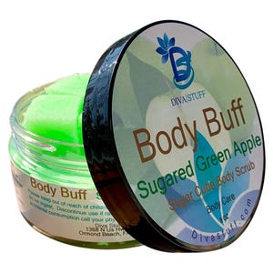 Puede incluir: Un tarro abierto de exfoliante corporal en cubos de azúcar "Body Buff", con aroma a "Manzana Verde Azucarada". El tarro muestra cubos de color verde brillante. La tapa muestra el logotipo de Diva Stuff e información del producto. El tarro contiene 227 gramos.