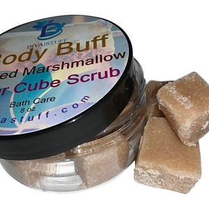 Puede incluir: Un tarro transparente de exfoliante de azúcar en cubos con tapa negra. La etiqueta dice "Body Buff Toasted Marshmallow Sugar Cube Scrub" y "Bath Care 8 oz". Varios cubos de azúcar moreno están junto al tarro.