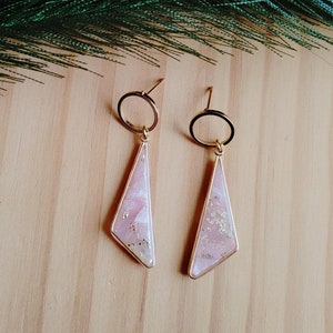 May include: A pair of gold-colored earrings with a geometric design. Each earring features a pink, pearlescent, and gold-flecked pendant suspended from a gold hoop. The earrings are displayed on a light wood surface, with a sprig of greenery in the background.