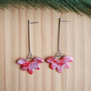 May include: A pair of dangle earrings with a floral design. The earrings feature a long, silver bar with a red, white, and pink marbled flower-shaped charm. The earrings are displayed on a wooden surface.