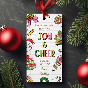 May include: A rectangular gift tag with a red ribbon, featuring colorful illustrations and text. The tag says "Thank You for Bringing Joy & Cheer to School This Year! Love, Hadley." Decorative elements include a school bus, apple, and pencil.