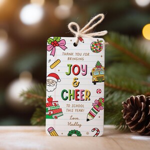 May include: A rectangular gift tag with a string, featuring colorful illustrations and text. The tag says "THANK YOU FOR BRINGING JOY & CHEER TO SCHOOL THIS YEAR! Love, Hadley." Illustrations include a school bus, Santa, and a stack of books.