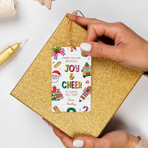 May include: A rectangular gift tag with the words "JOY & CHEER" and school-themed illustrations, held over a gold glitter gift box. The tag includes the text "THANK YOU FOR BRINGING TO SCHOOL THIS YEAR! Love, Hadley."